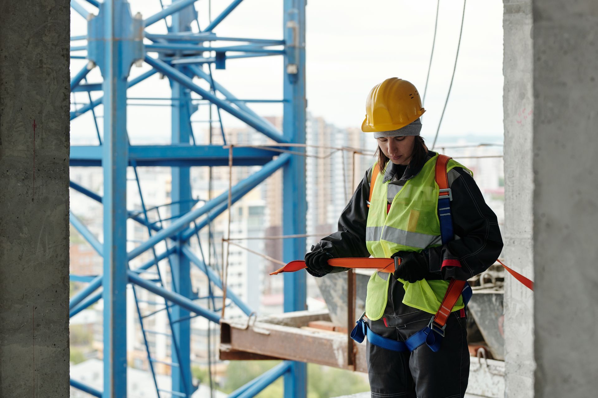 Construction worker in hard hat and safety vest, attaching harness to a beam, crane in the background.