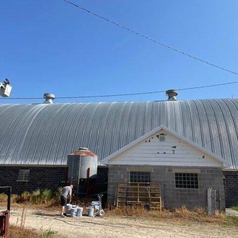 White barn with a curved metal roof, small front entry, and a person working beside equipment outside.