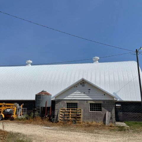 Farm building with a white metal roof, small gray entryway, and equipment beside it under a clear blue sky