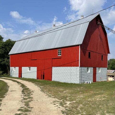 Large red barn with white lower walls and a gray metal roof beside a dirt path under a blue sky