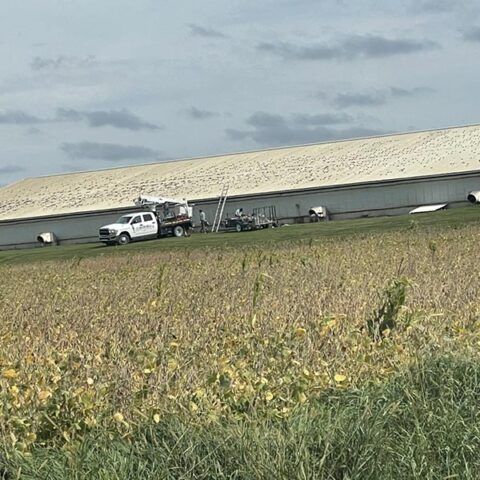 White truck and farm equipment beside a long metal building with a pale roof in a grassy field.