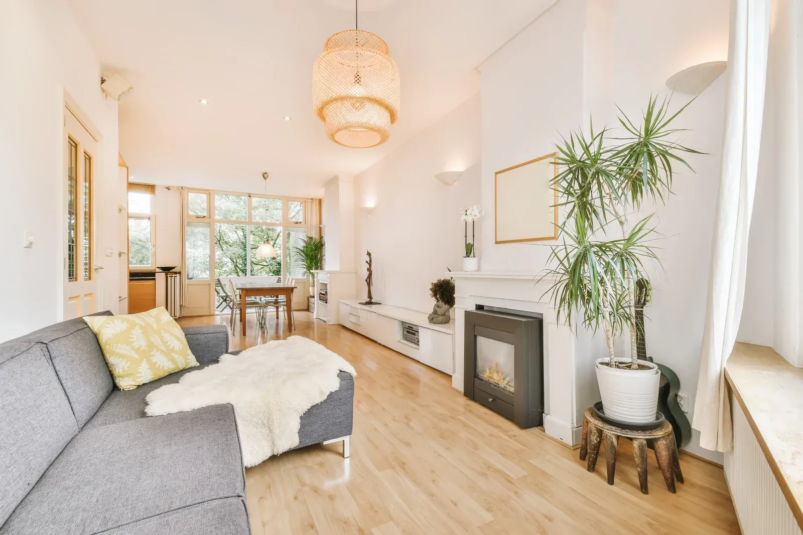 Bright modern living room with gray sectional sofa, wood floors, potted plant, and dining area beyond.