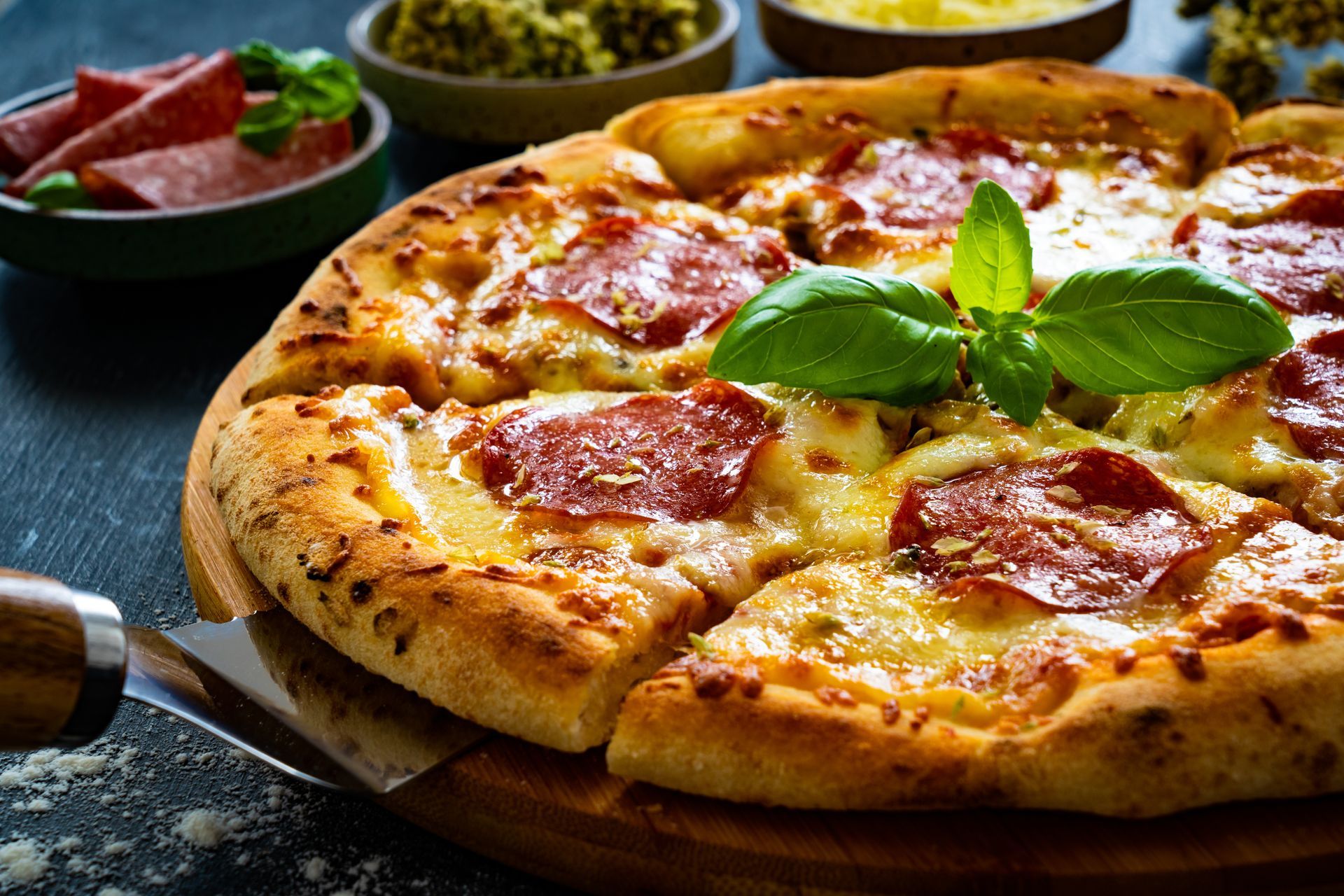 Pepperoni pizza with basil on a wooden board, with bowls of toppings in the background