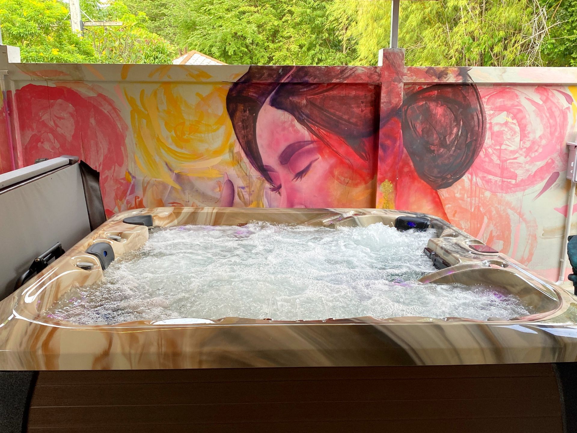 Outdoor hot tub in a side yard, water bubbling, surrounded by a colorful mural of a woman’s face on the privacy wall with green trees visible above.