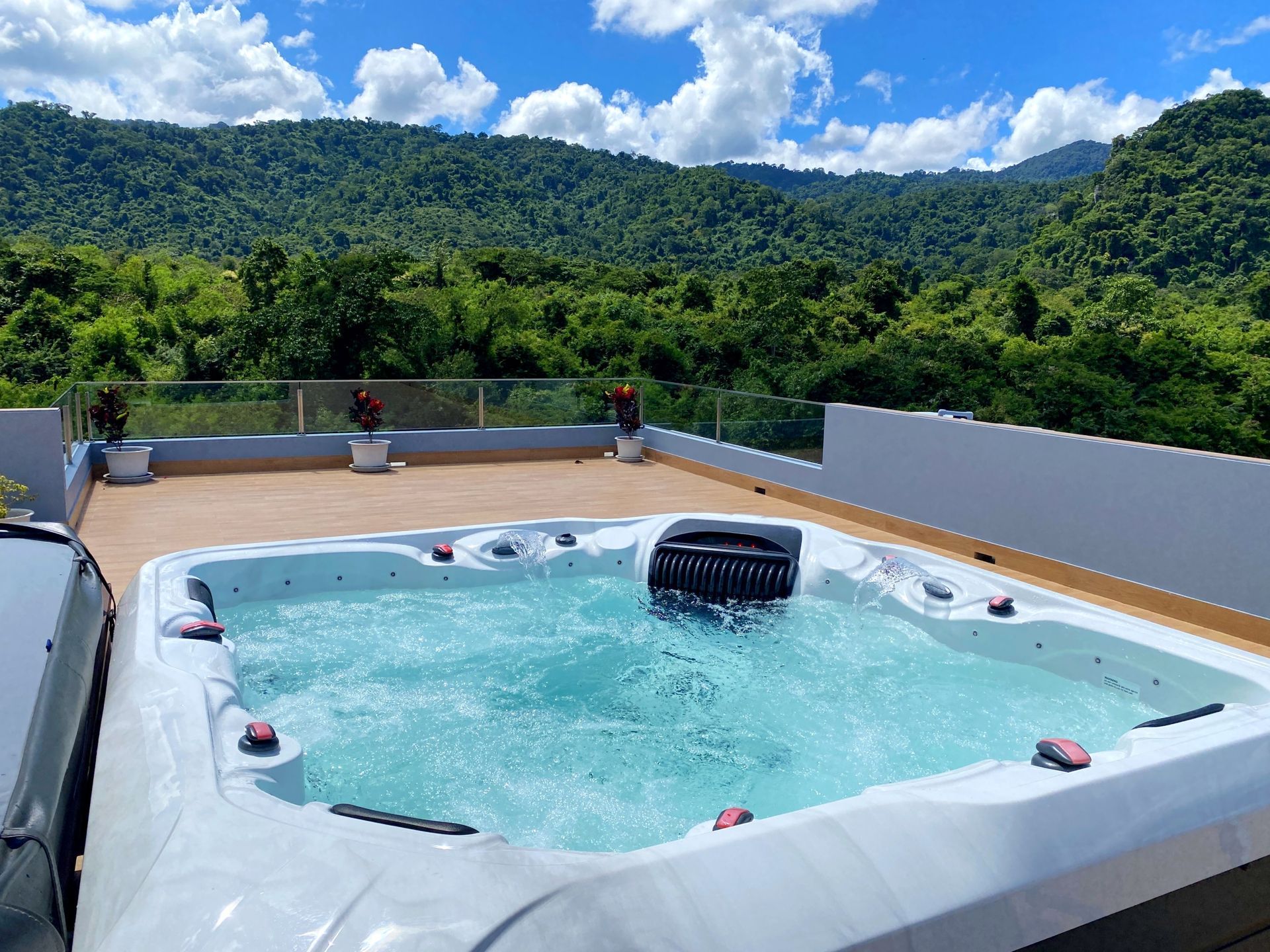 Large outdoor hot tub on a rooftop deck with glass railing, potted plants, and a lush green mountain
