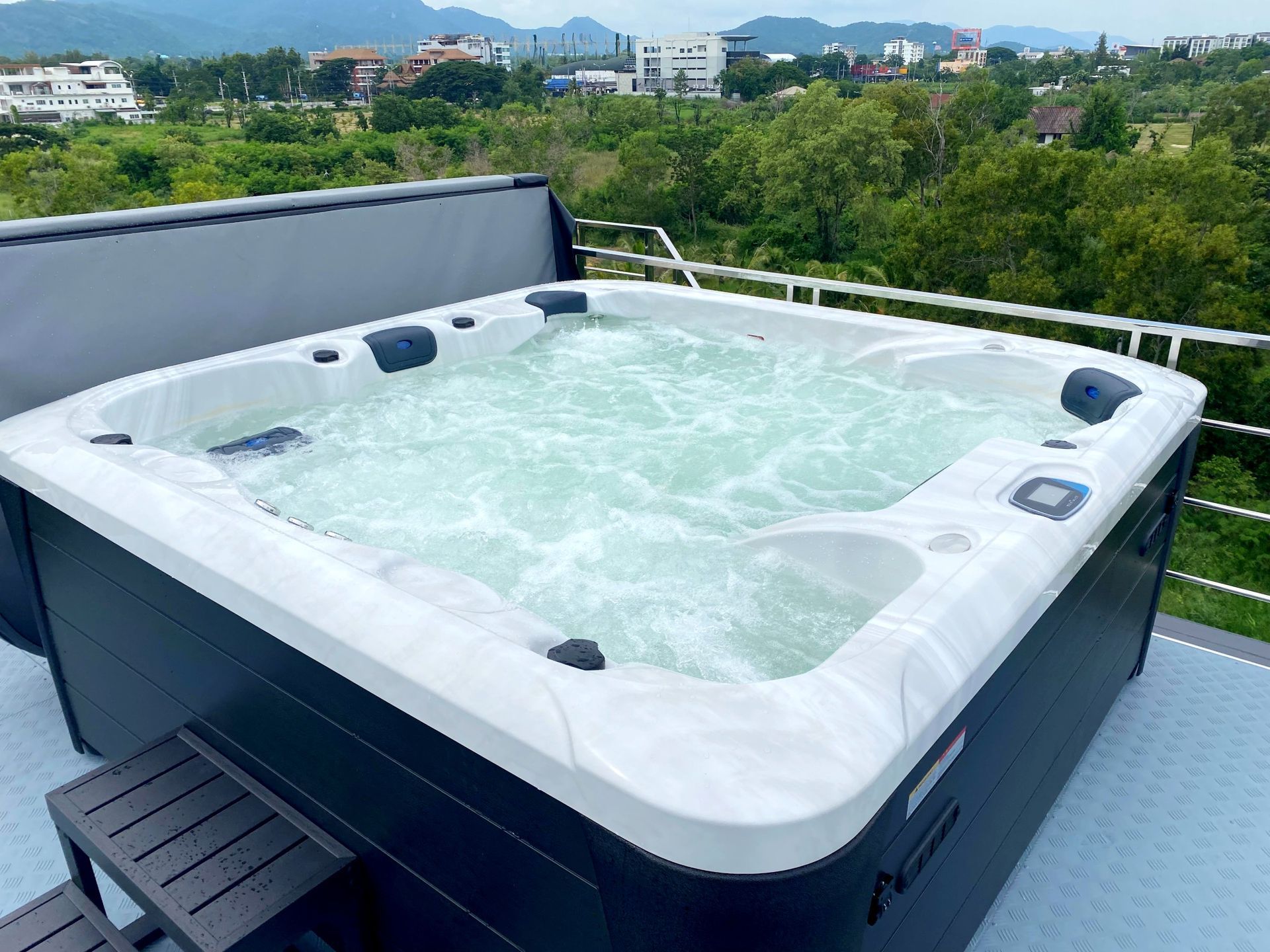 Large white outdoor hot tub on a rooftop deck with jets running, small step stool beside it, overlooking green trees, city buildings, and distant mountains.