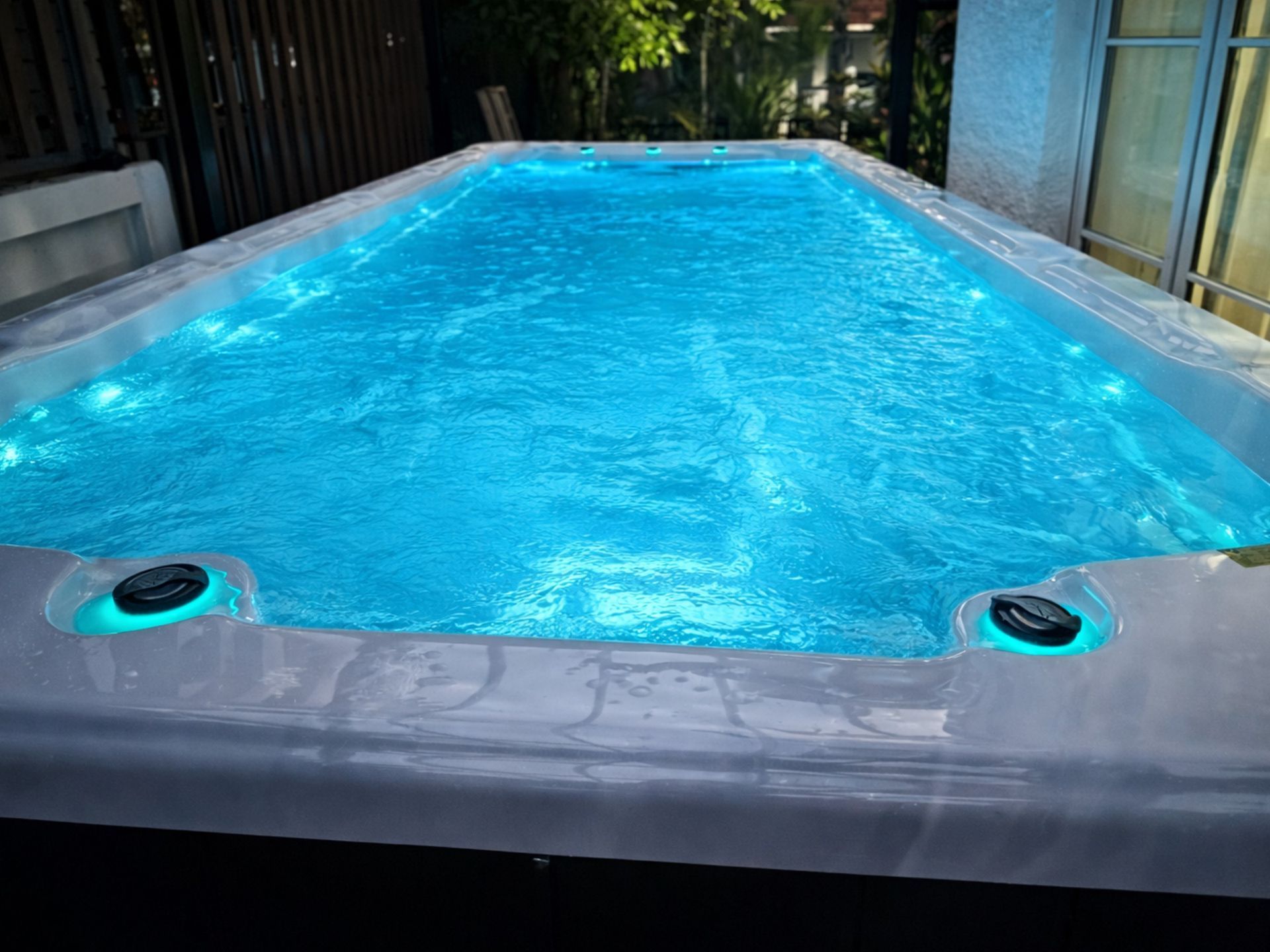 Outdoor swim spa filled with clear blue water and ambient LED lighting, installed in a residential garden area for swimming, exercise, and relaxation.