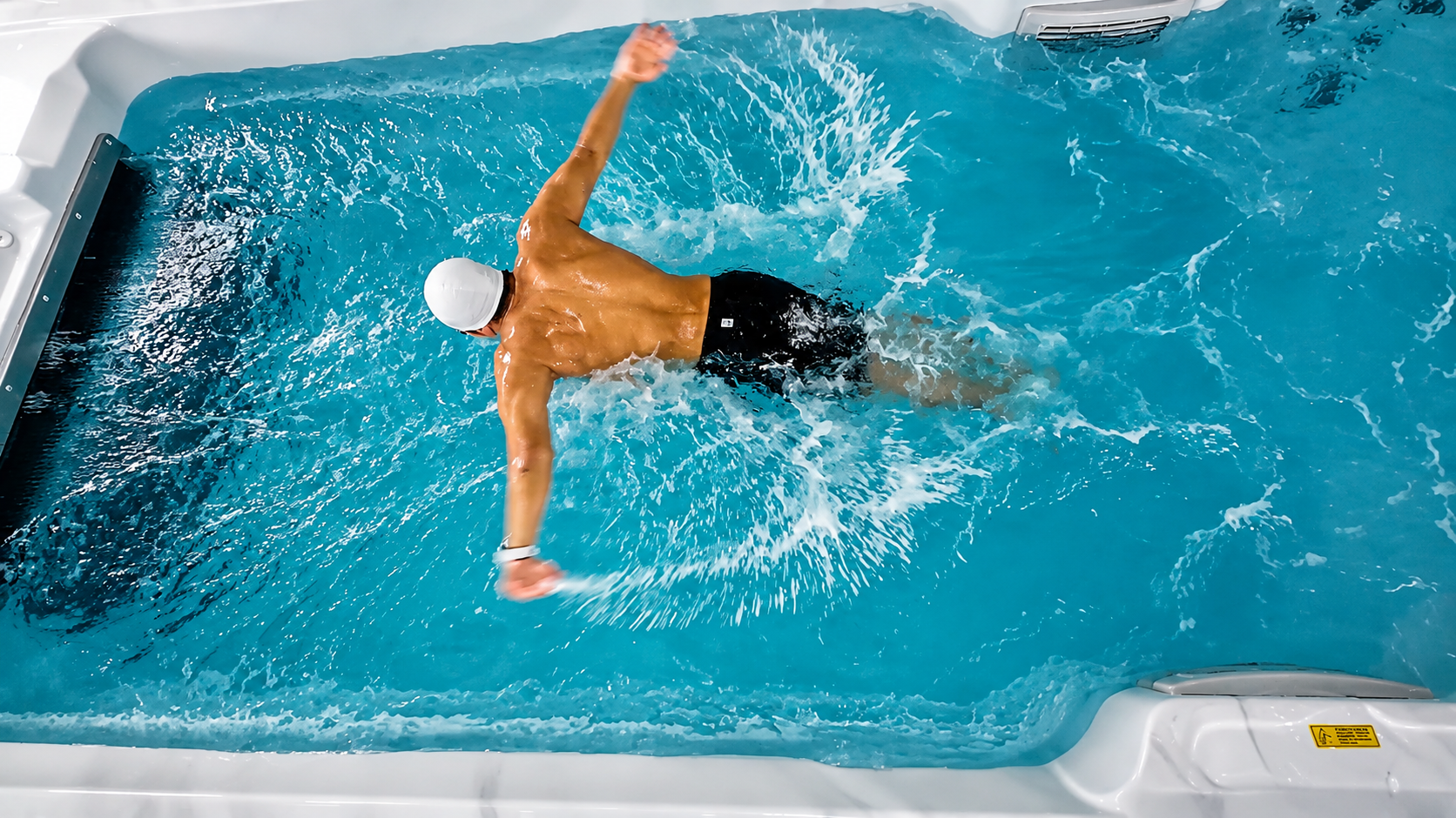 Top view of a swimmer training in a swim spa with powerful counter-current system, creating dynamic splashes and wave motion in clear blue water.