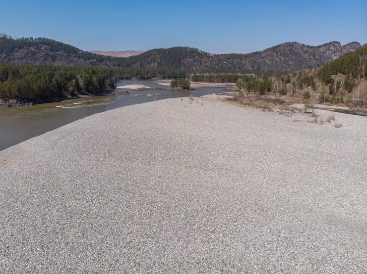 Dry riverbed with pale gravel, a narrow stream, and low forested mountains under a clear sky