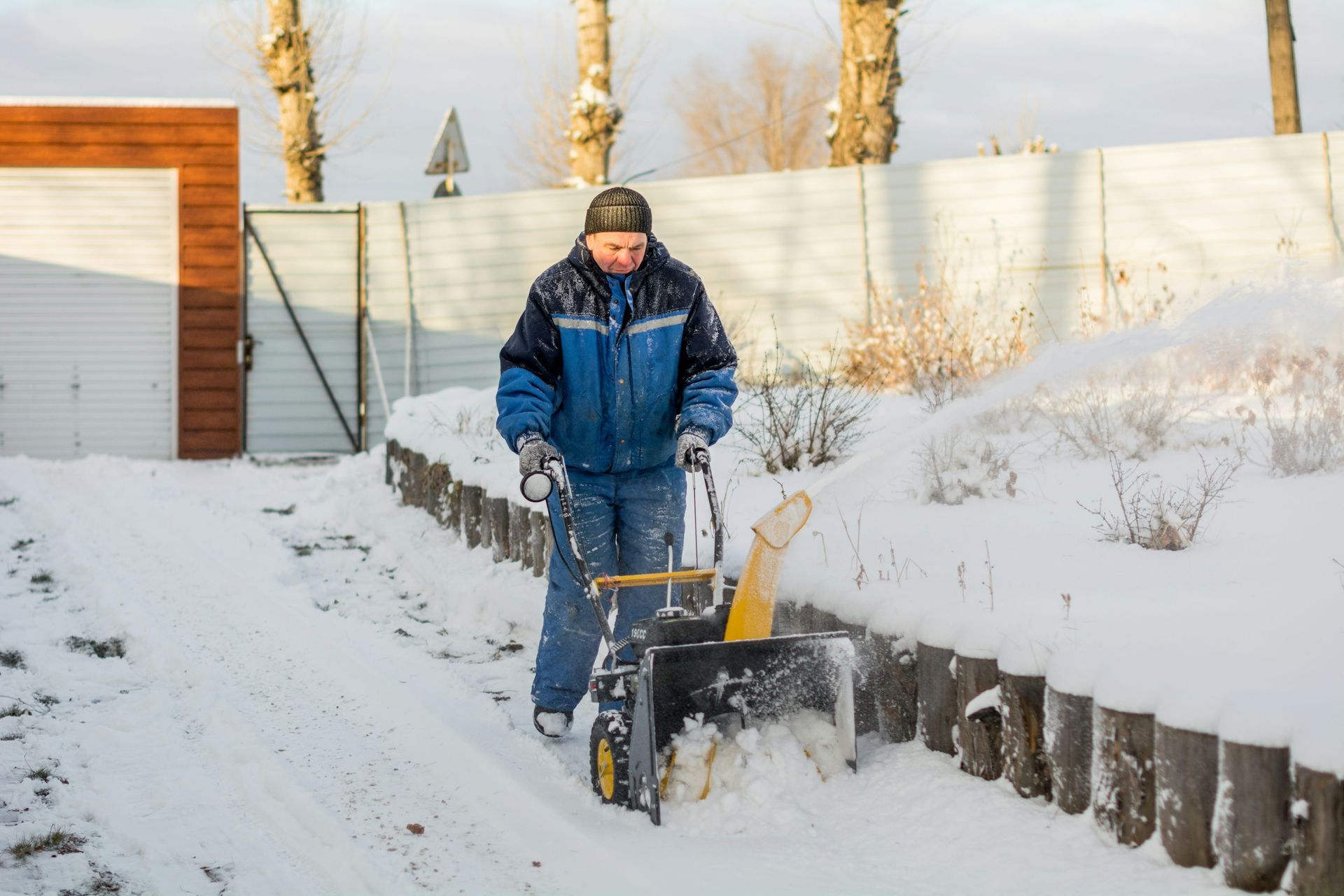Person shoveling snow in a snowy yard beside a fence and wooden shed