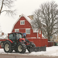 Red tractor plowing snow in front of a red farmhouse and bare trees