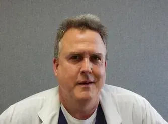 Man in a white coat against a gray background, facing the camera with a neutral expression