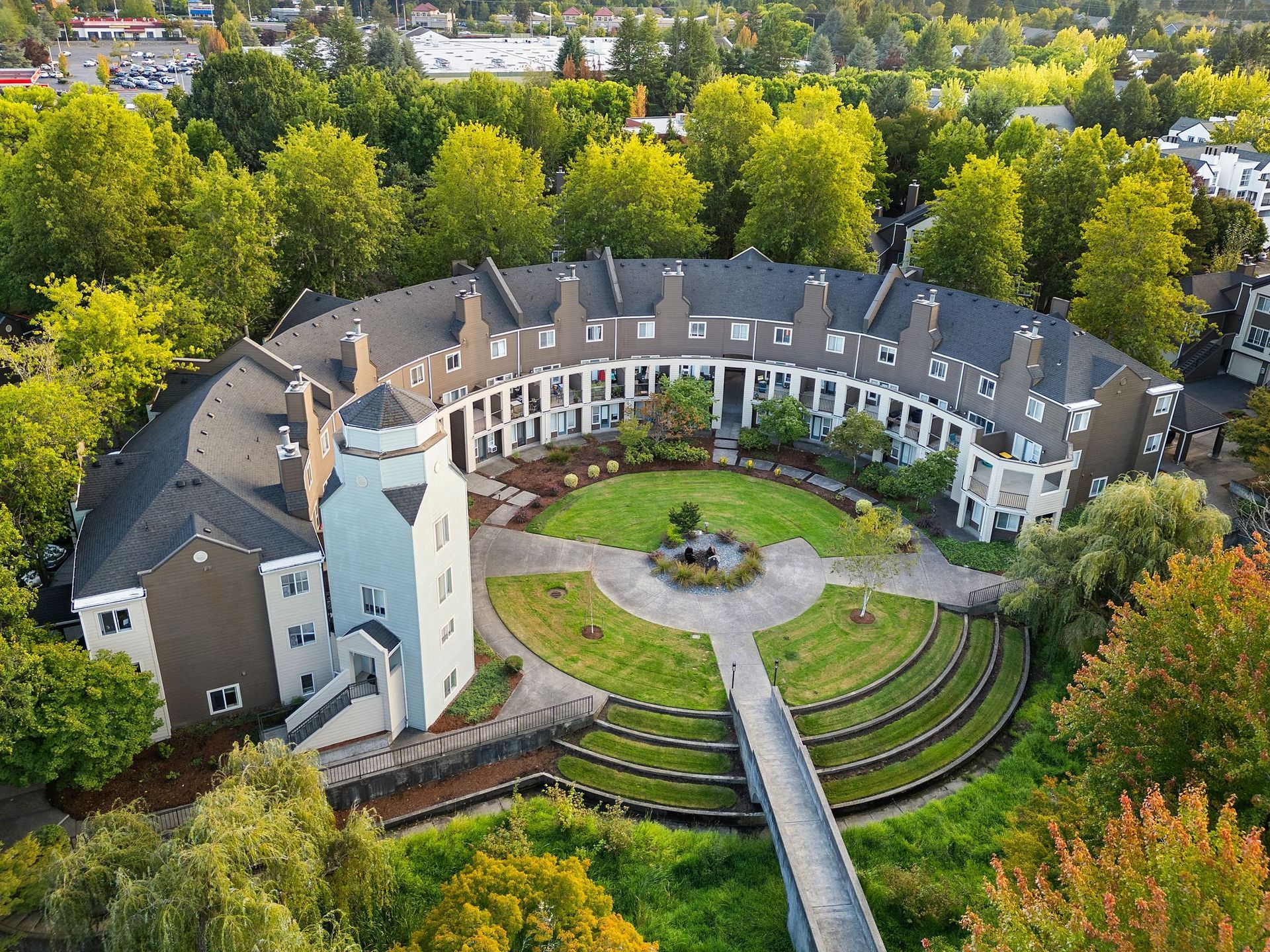 Aerial view of a curved apartment complex surrounding a central green courtyard. A blue-gray tower and a bridge are visible.