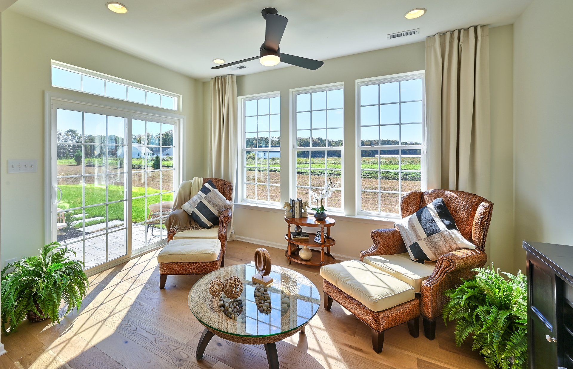 Bright sunroom with wicker chairs, white curtains, a ceiling fan, and views of a green garden.
