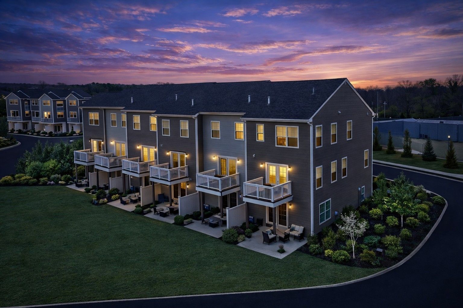 Row of townhouses at dusk with lit windows and patios beside a green lawn under a purple sunset sky