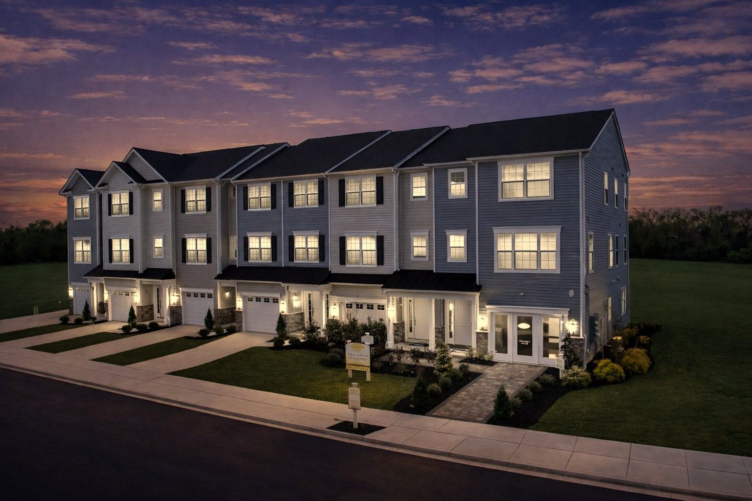 Row of illuminated townhouses at dusk with lit windows, sidewalks, and landscaped front lawns