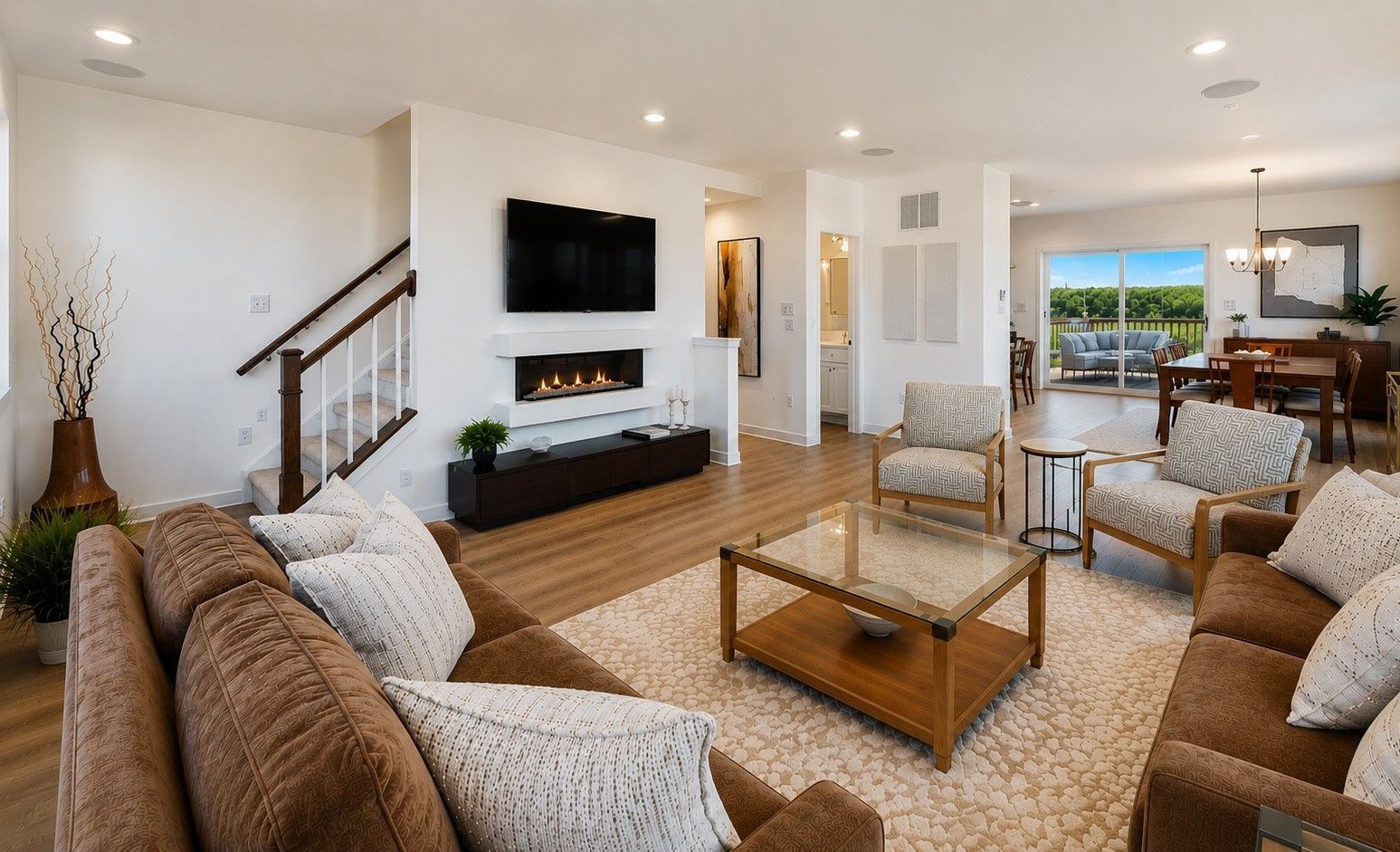 Bright living room with tan sofa, fireplace TV wall, coffee table, staircase, and large windows showing greenery