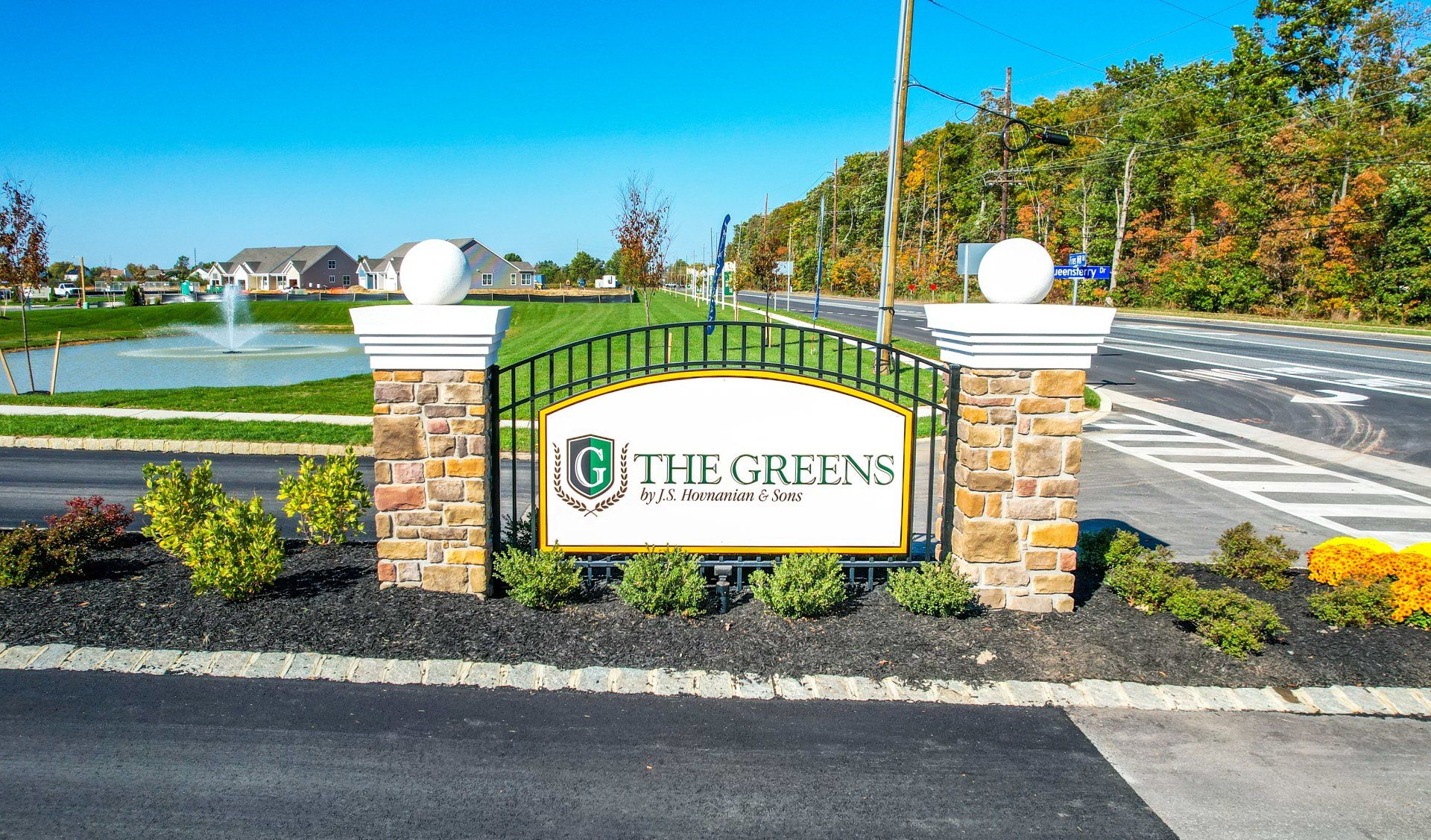 Entrance sign for The Greens community with stone pillars, round lights, and landscaped roadway