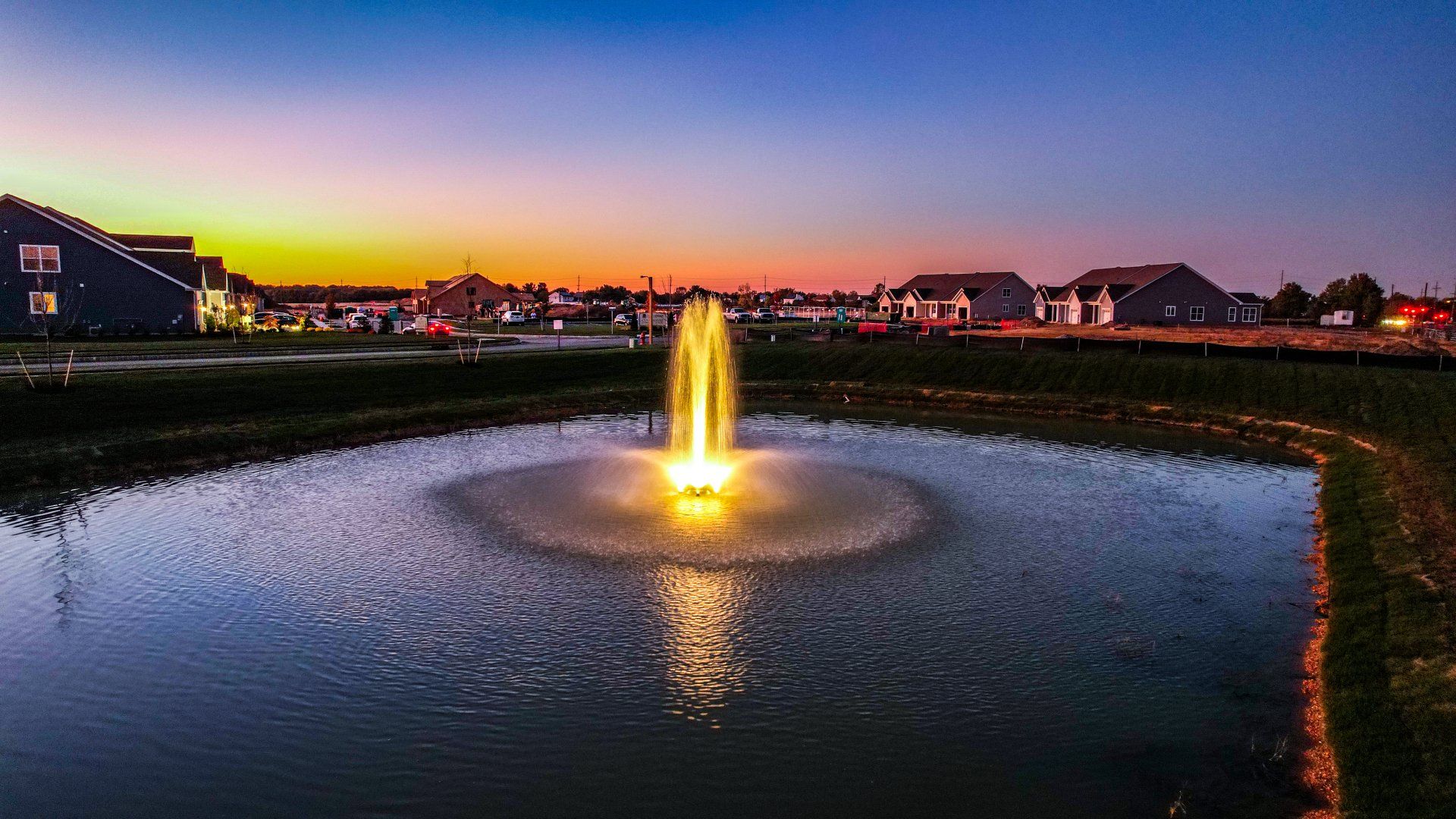 A lit fountain in a pond at dusk with houses and colorful sunset sky in the background