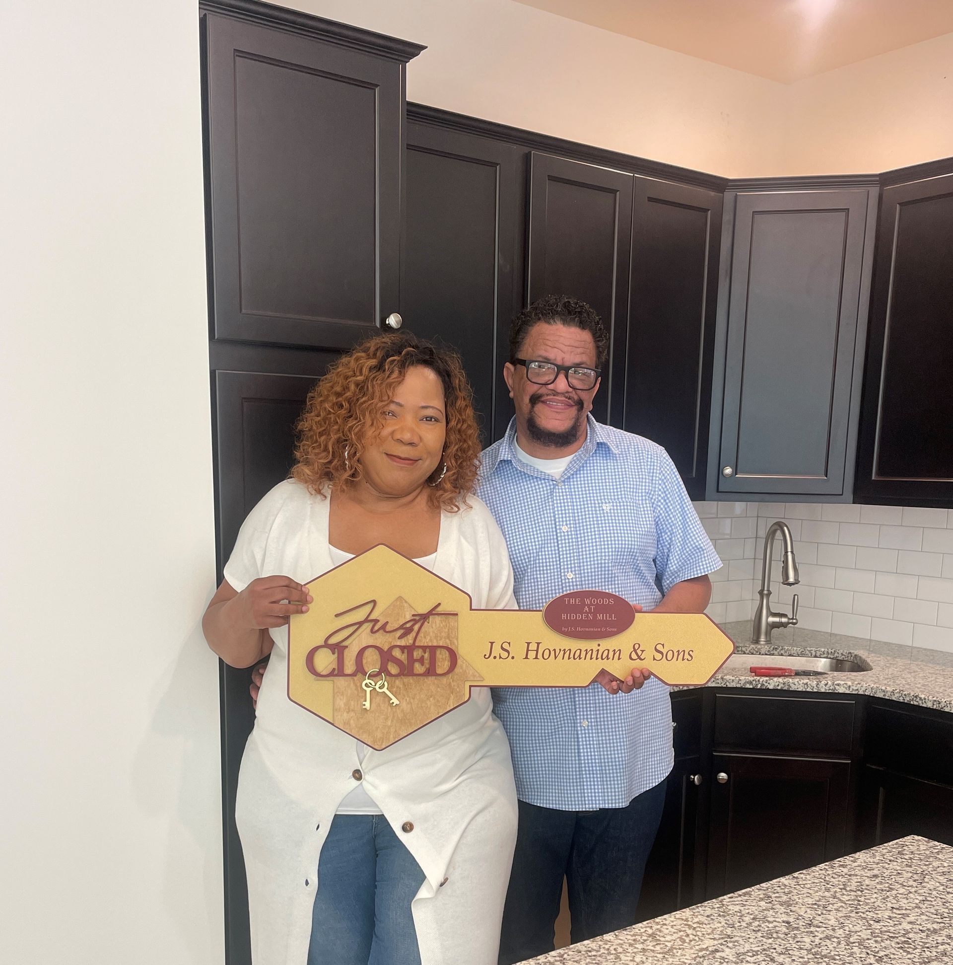 Two people in a kitchen holding green “Closed” and “U.S. Home… & Sons” signs