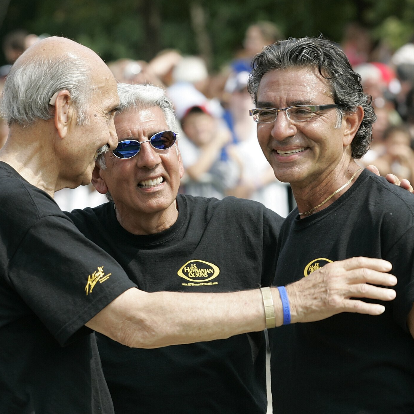 Three smiling people in black shirts embracing outdoors at a gathering.