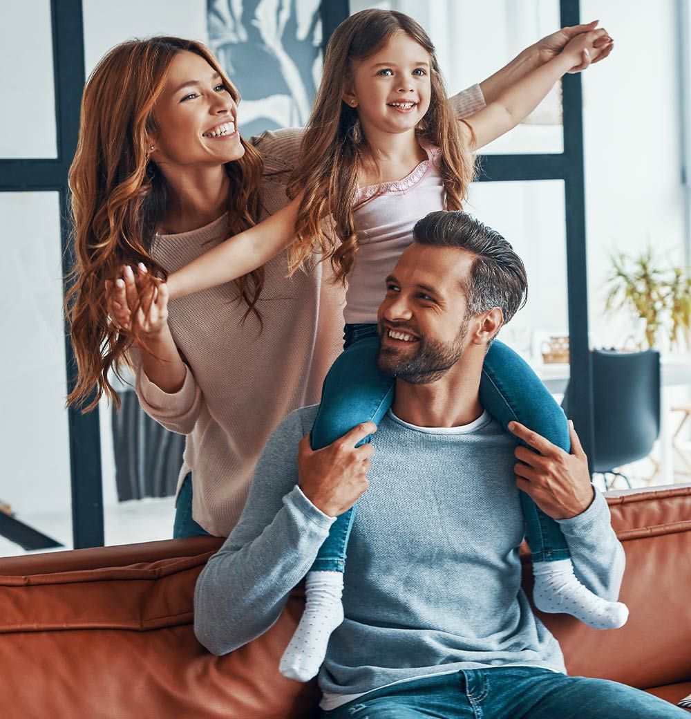 Smiling family indoors, with two children on an adult’s shoulders in a bright living room.