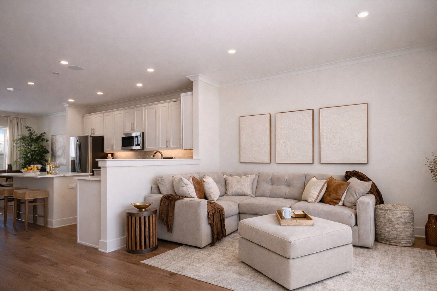 Bright, neutral living room with beige sofa, ottoman, wall art, and open kitchen in the background