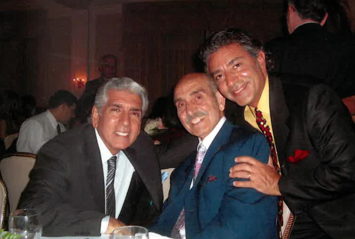 Three men in suits smiling at a formal dinner table with dim, warm lighting.