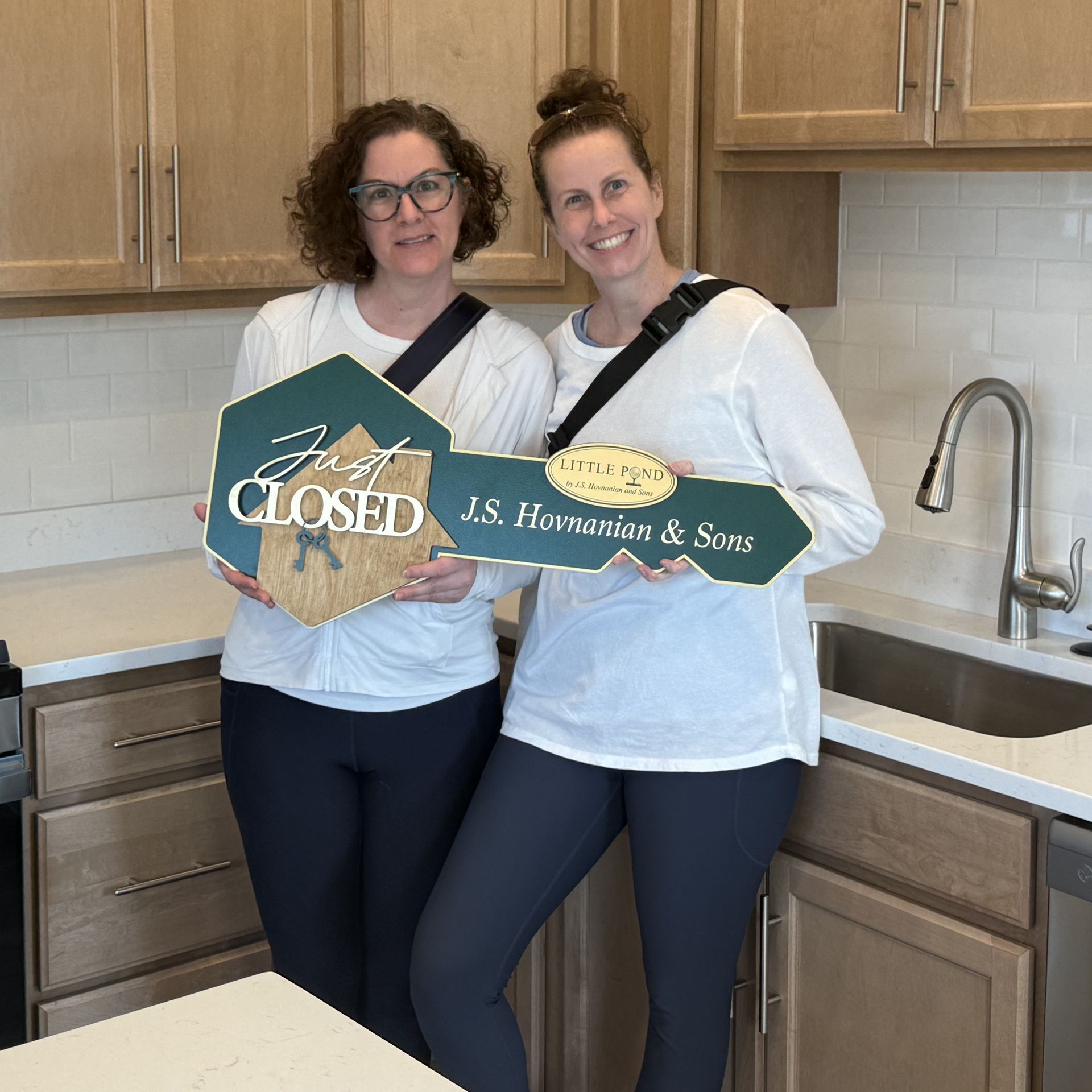Two women in a kitchen holding a “CLOSED” sign and a house-shaped sign.