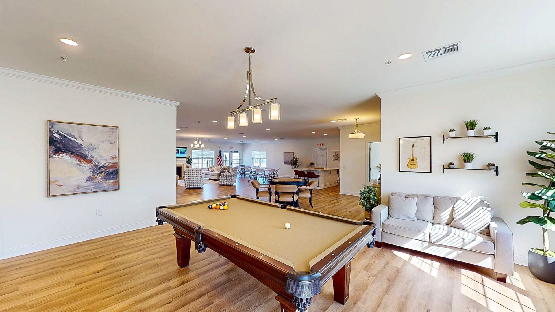 Bright living room with pool table, white sofa, wood floor, and open kitchen in the background