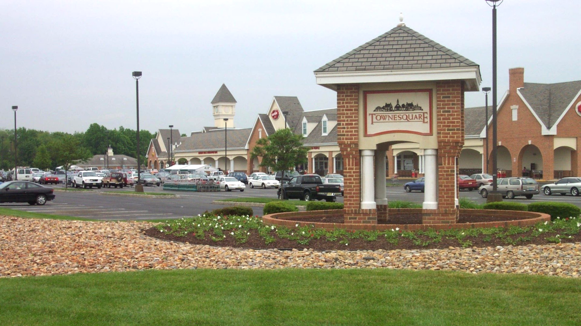 Brick entrance sign in a landscaped parking lot with cars and a building in the background.