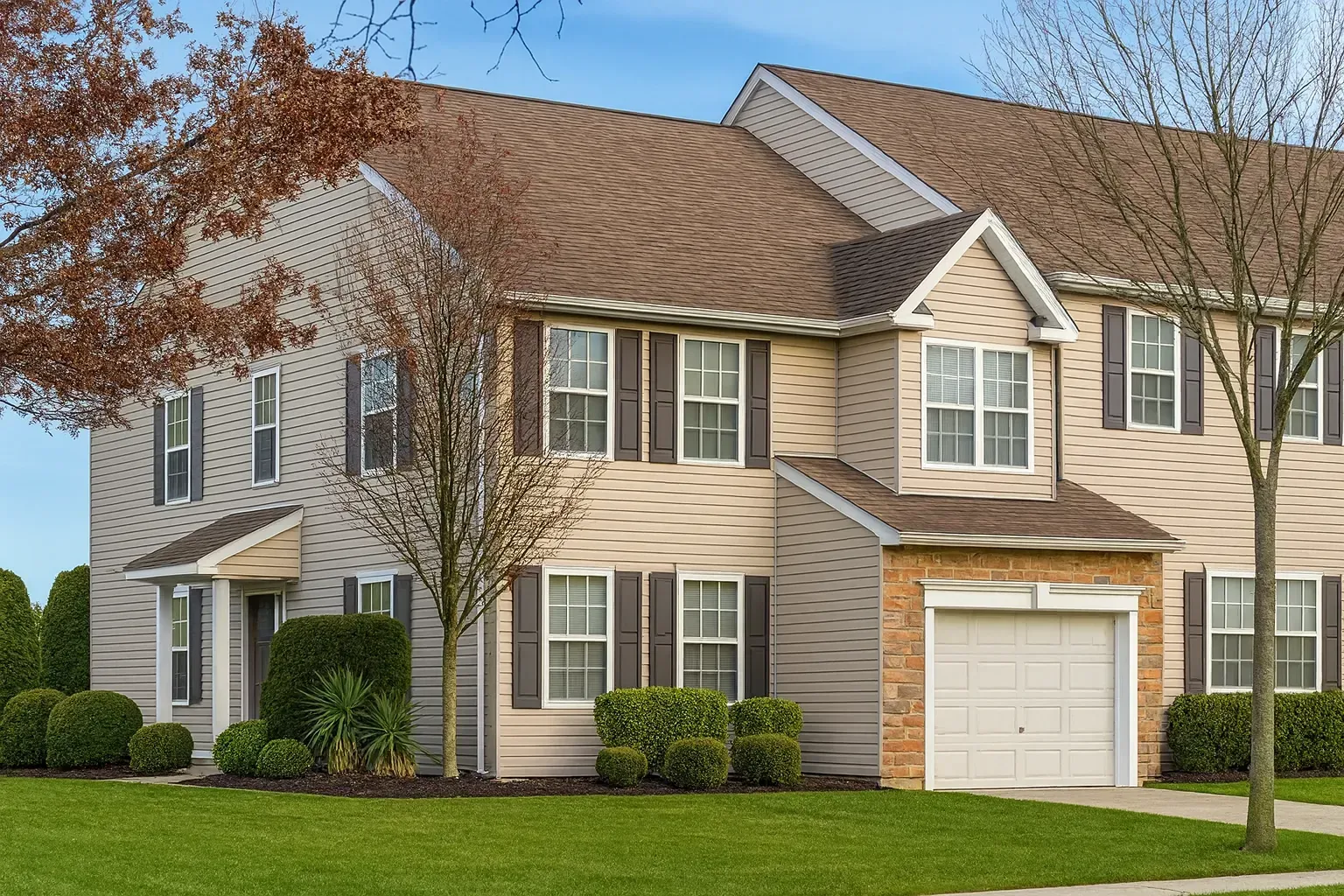 Suburban beige townhouse with white garage, green lawn, shrubs, and leafless tree under blue sky
