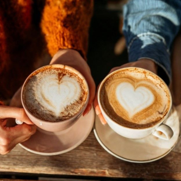 Two cups of latte with heart-shaped foam on a wooden table, held by two hands