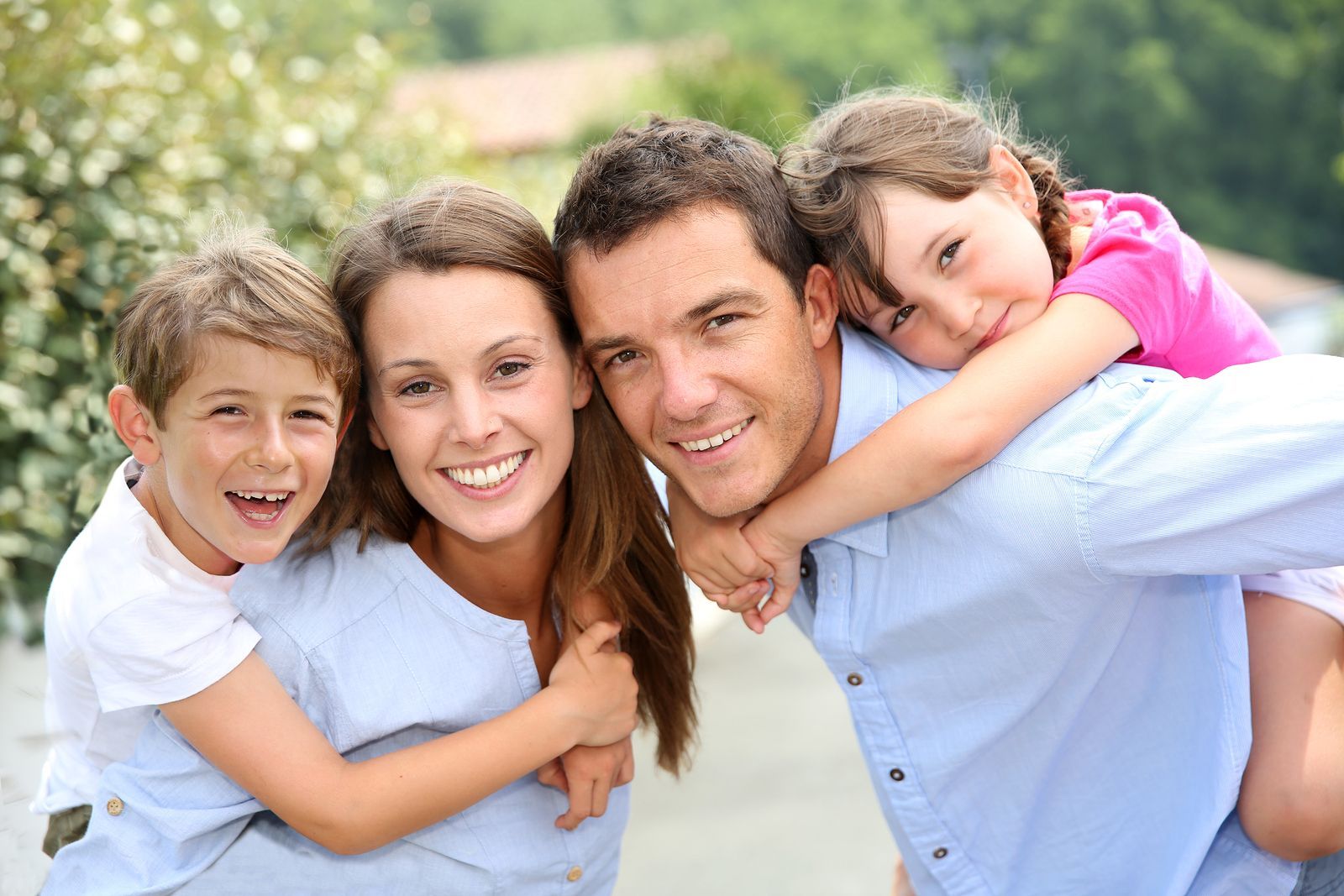 Smiling family of four outdoors, with parents carrying two children on their backs.
