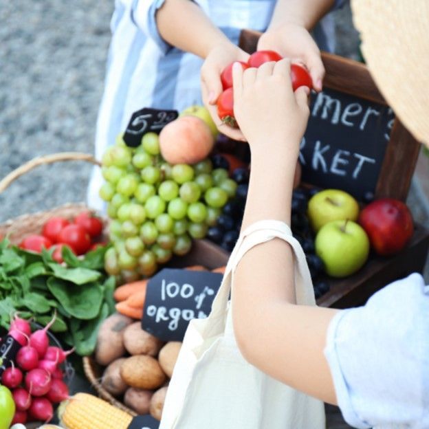 Hands arranging fruit and vegetables at an outdoor market stall with handwritten price signs