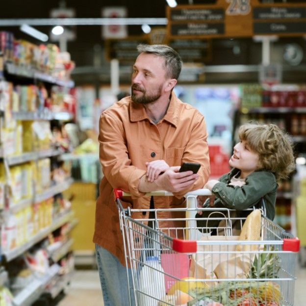 Man shopping with a child in a grocery store, pushing a cart and checking a phone.