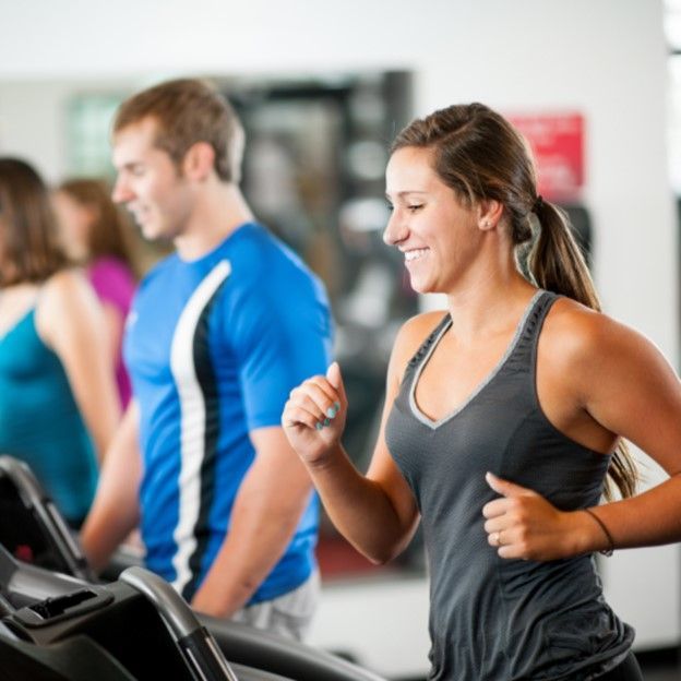 People exercising on treadmills in a bright gym, with a woman in a gray tank top smiling in the foreground