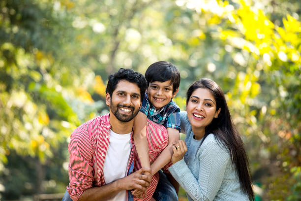 Smiling family of three outdoors, with a child on each parent’s back in a sunlit garden.