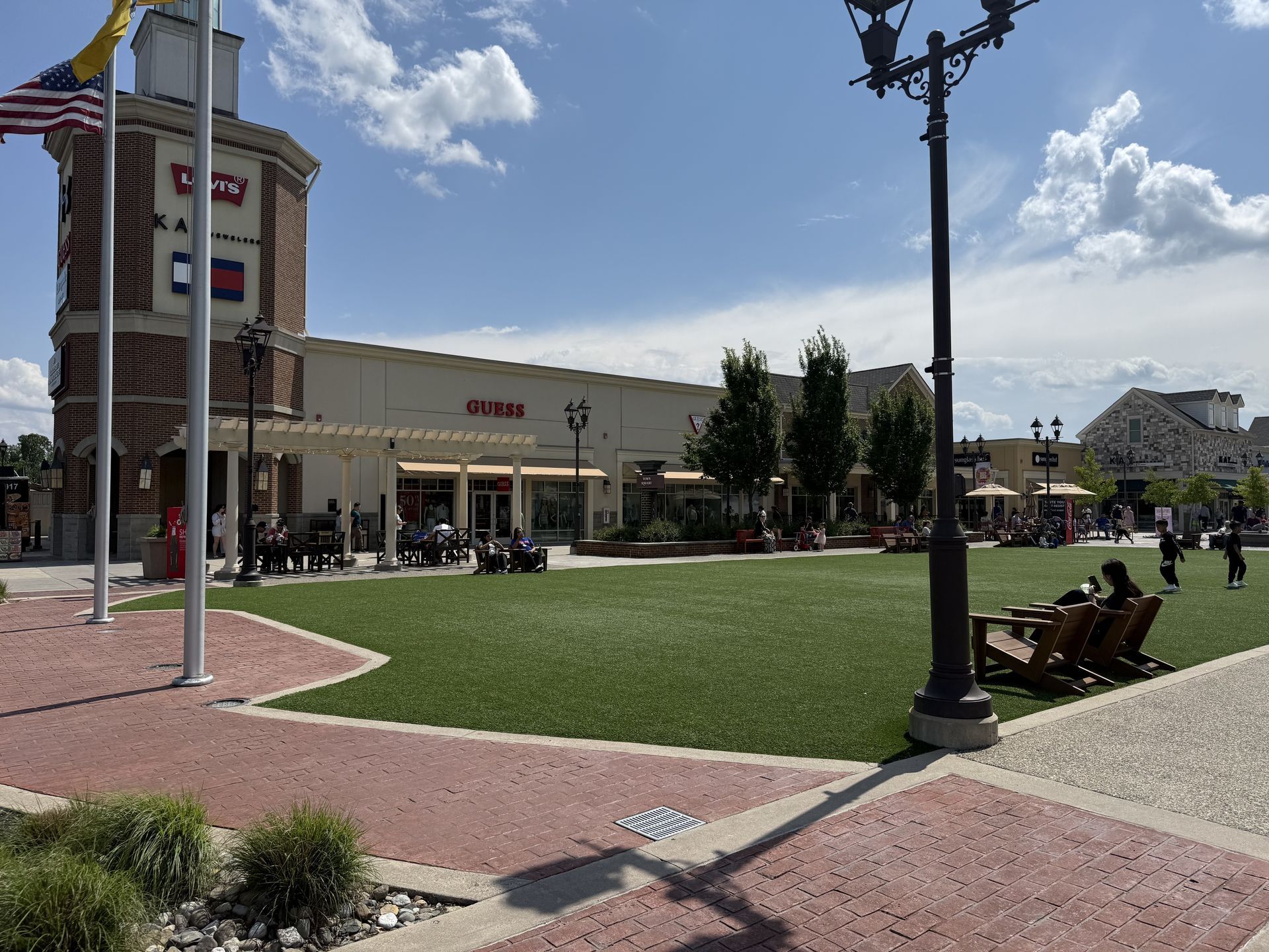Outdoor shopping plaza with green lawn, brick paths, storefronts, lampposts, and people under a blue sky
