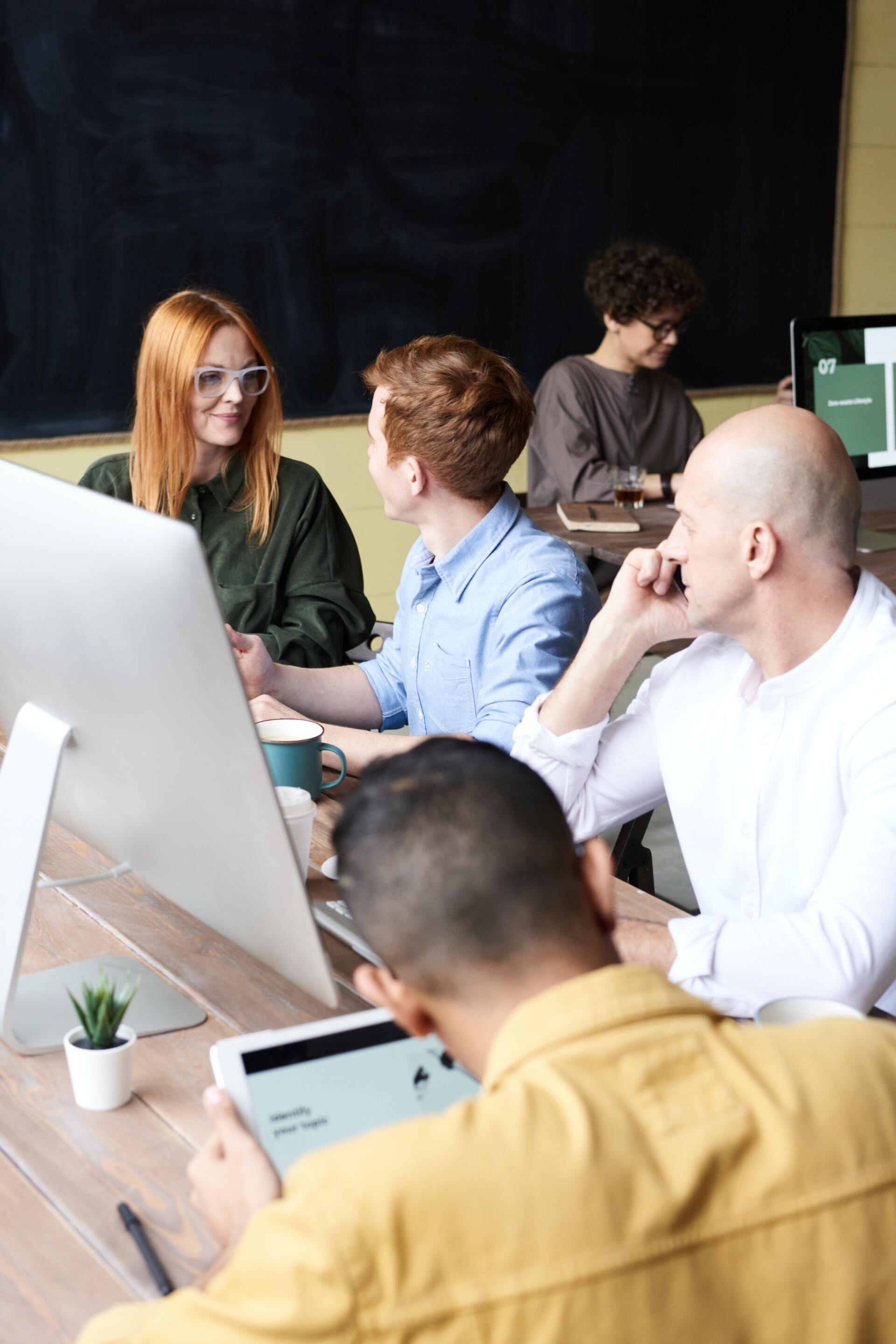 Colleagues in a meeting around a desk, discussing work in an office with laptops and monitors.