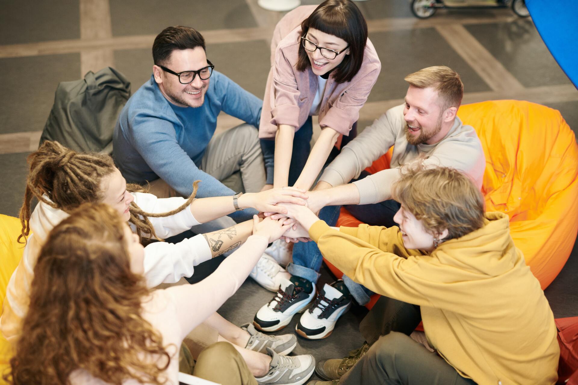 Group of people in a circle stacking hands together in a team huddle, smiling indoors.