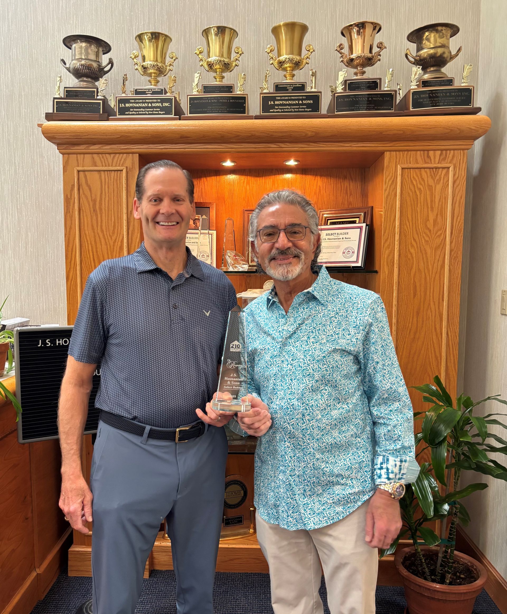 Two men posing with a trophy in front of a wooden cabinet topped with awards.