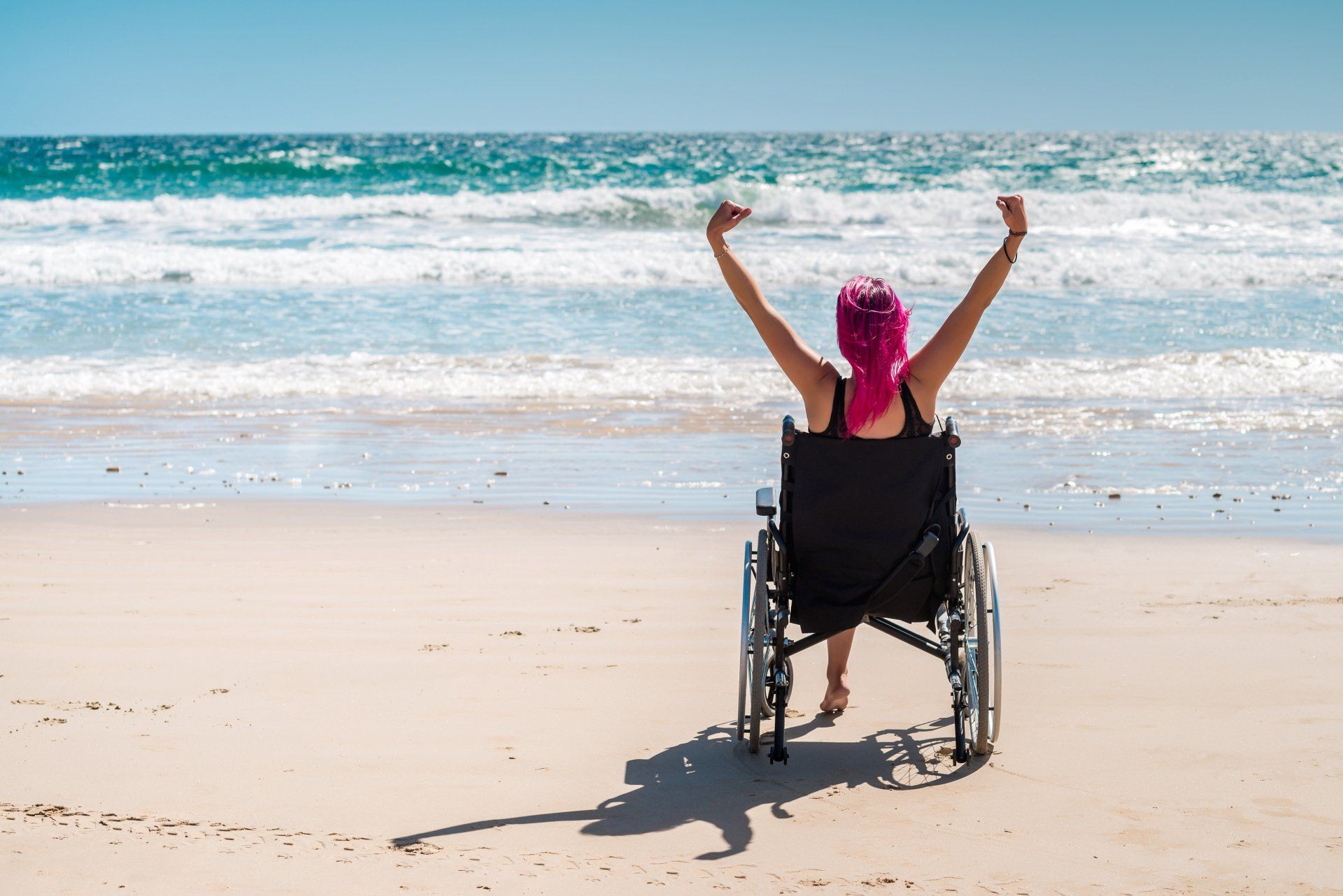 A woman in a wheelchair is sitting on the beach with her arms in the air.