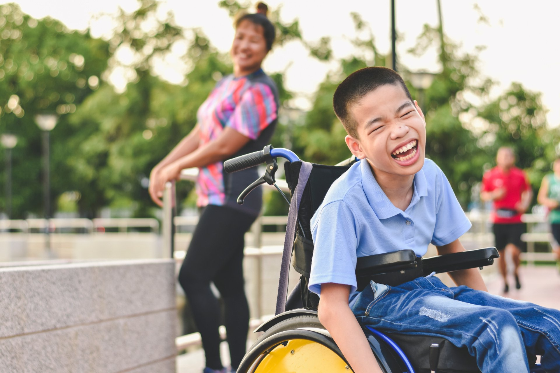 A young boy in a wheelchair is smiling while a woman stands behind him.