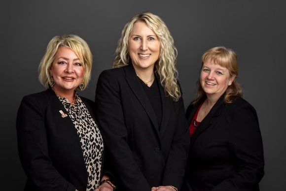 Three women in business attire, posing. Dark blazers, smiling, against a gray backdrop. Three women in business attire, posing. Dark blazers, smiling, against a gray backdrop.