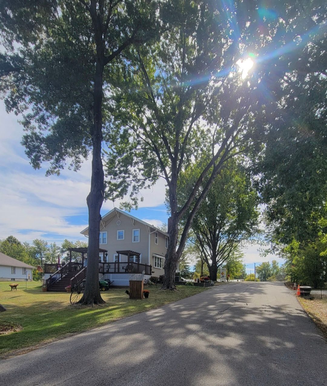 Tree-lined residential street with a house, parked car, and bright sun shining through the trees.