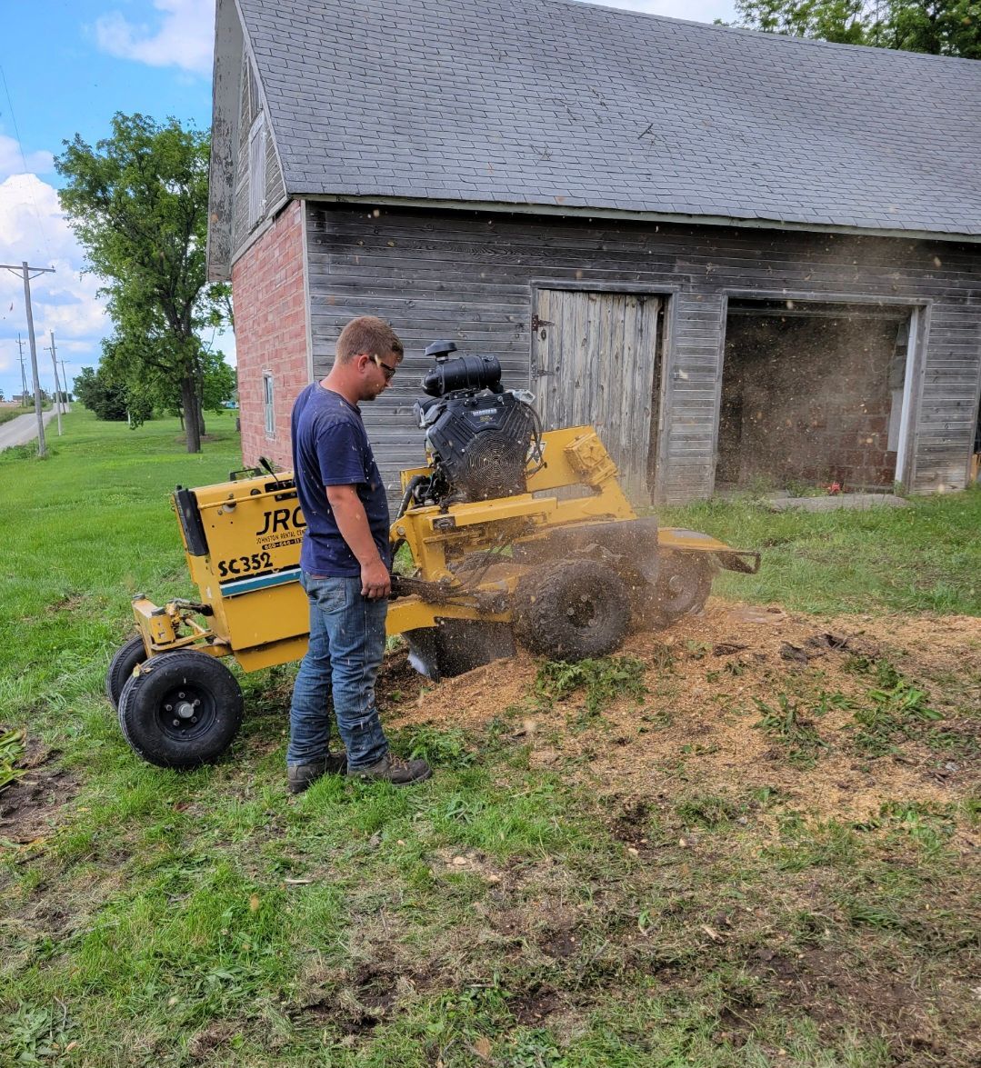 Man using a yellow stump grinder beside a barn, grinding dirt and debris into the grass.