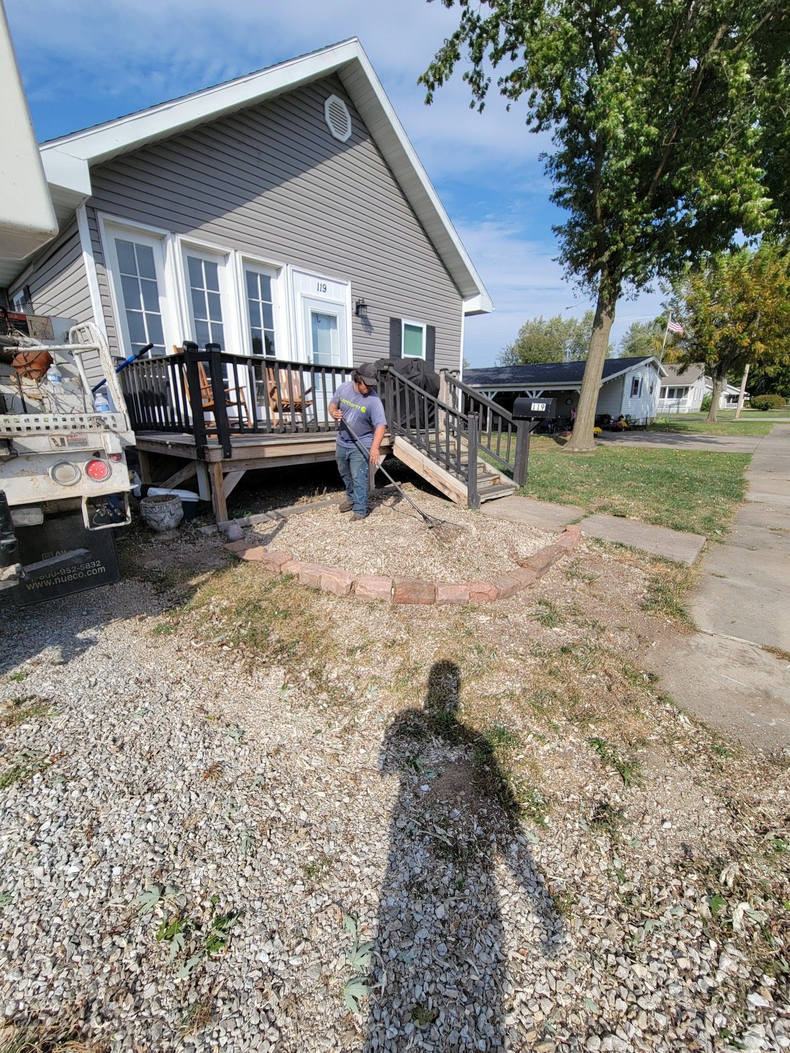 Person standing by a gray house with a porch and gravel yard on a sunny day
