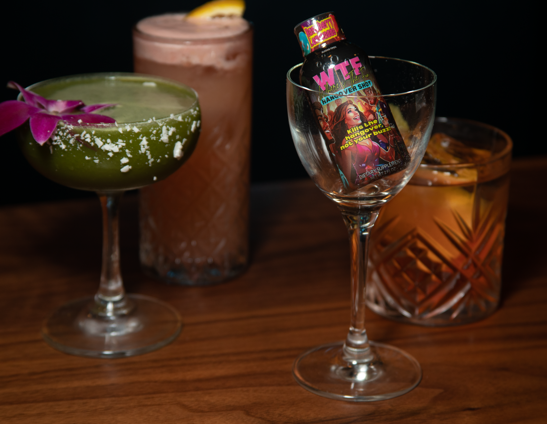 Four varied cocktails on a wooden surface, including a green drink with a floral garnish and a mini bottle in a glass.