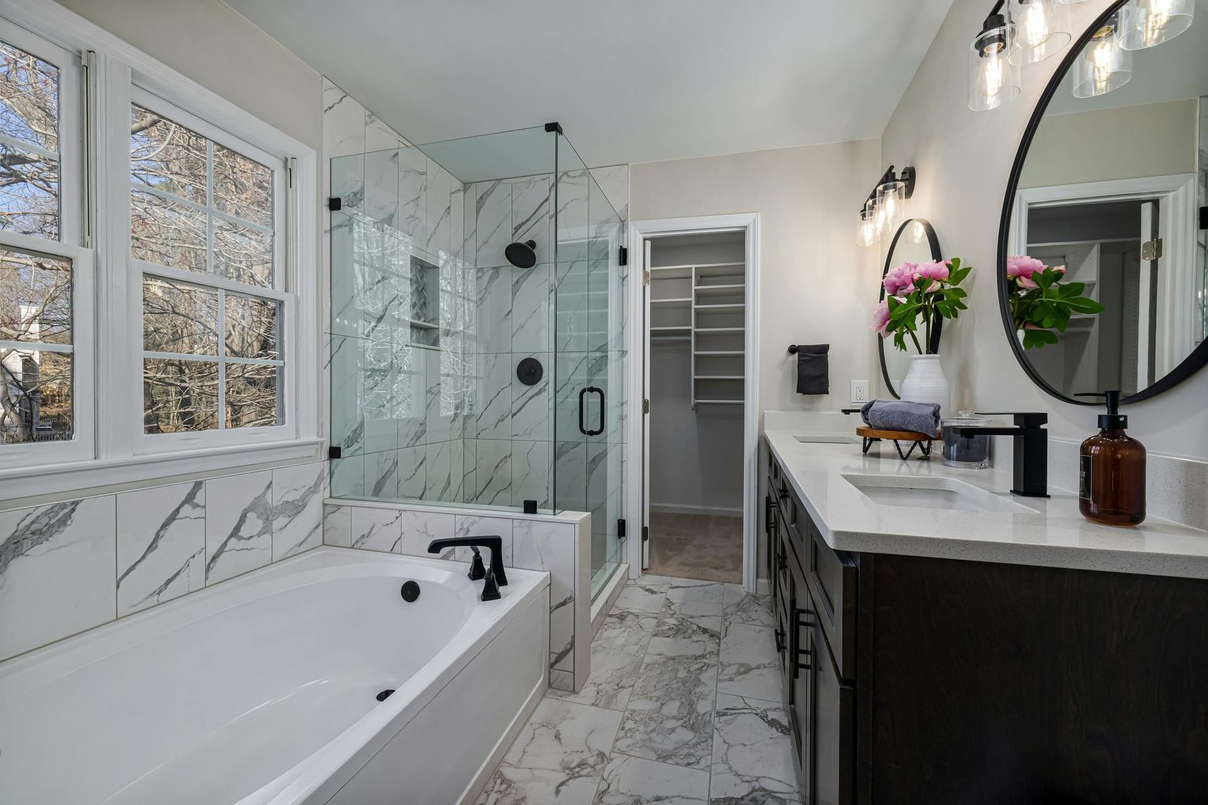 Modern white bathroom with soaking tub, glass shower, marble tile, and vanity with round mirror.